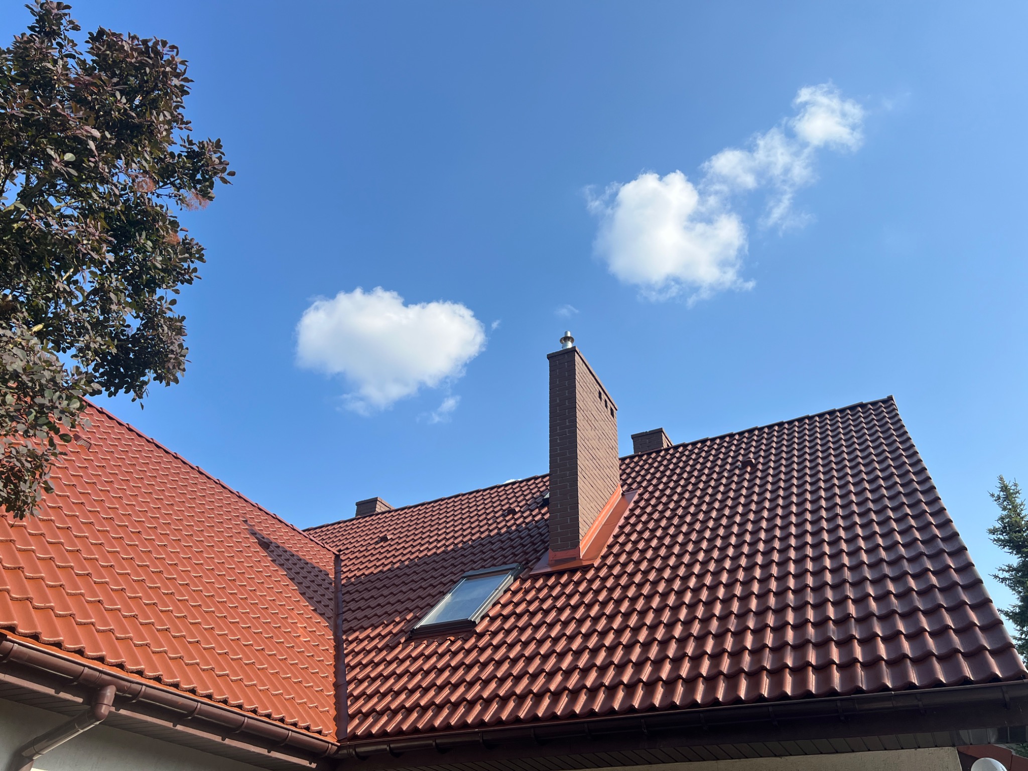 Czerwony dach z dachówki ceramicznej i ceglany komin na tle błękitnego nieba z chmurami. Widoczne okno dachowe i fragment drzewa.