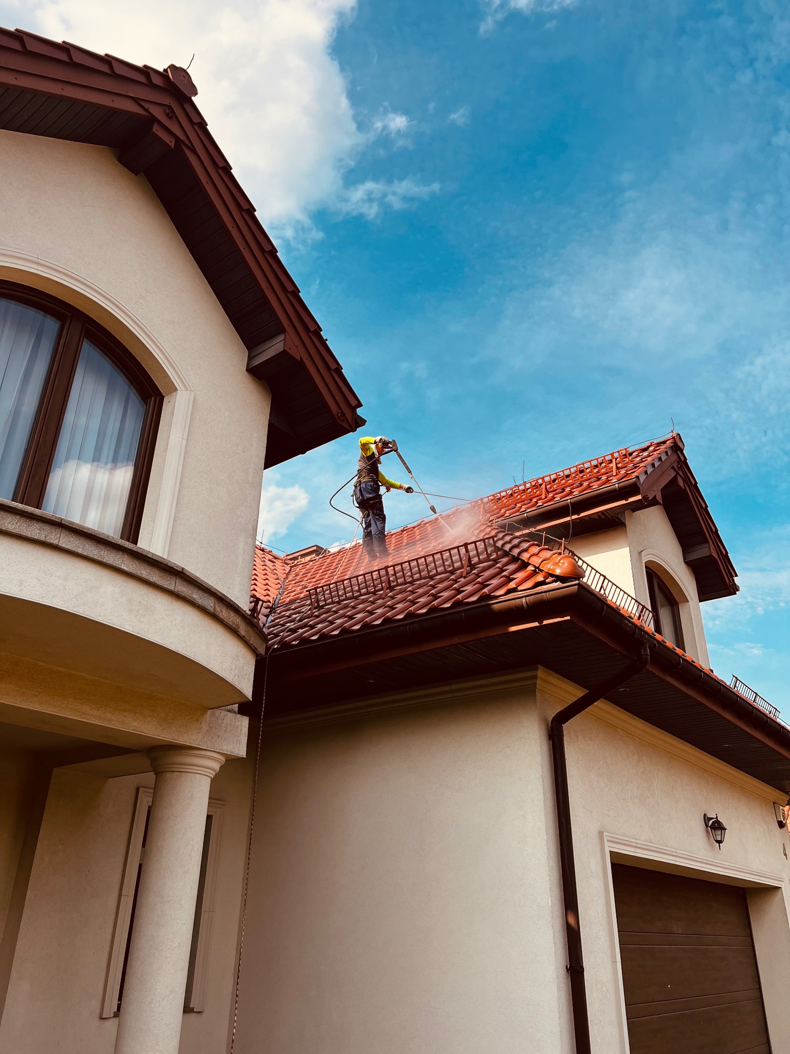 Pracownik w uprzęży czyści czerwoną dachówkę domu jednorodzinnego za pomocą myjki ciśnieniowej. Widok od dołu na tle błękitnego nieba.
