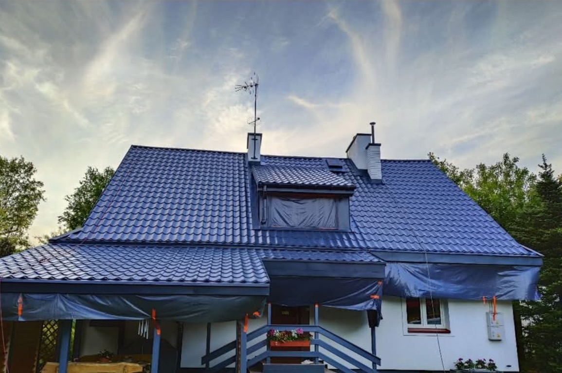 Dom z ciemnoniebieskim dachem krytym blachodachówką, zabezpieczony folią malarską, z anteną na kominie i kwiatami na ganku. Widok na elewację i fragment ogrodu.