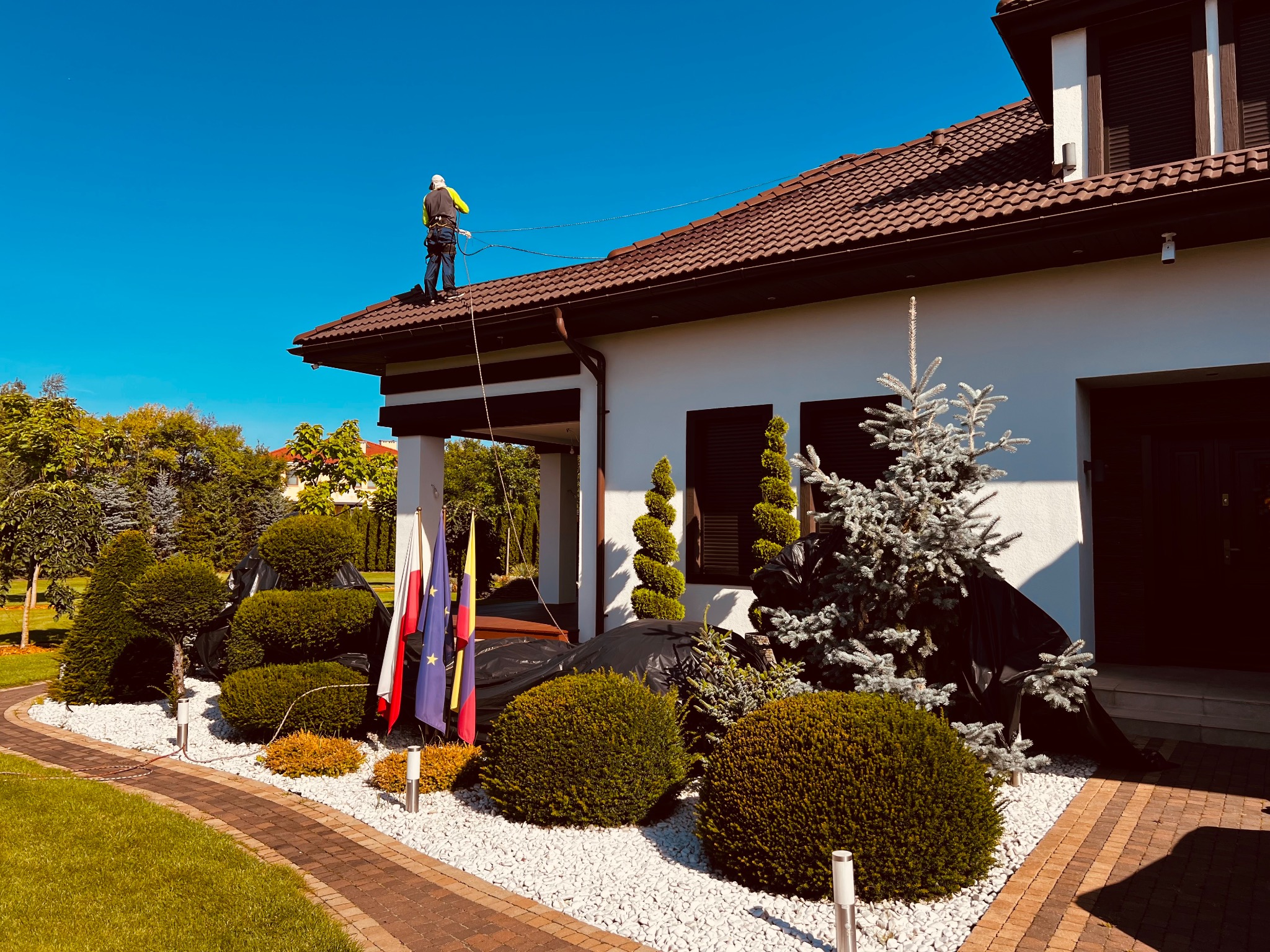 Pracownik w uprzęży na brązowym dachu domu podczas prac. Widoczne flagi i ozdobne krzewy w otoczeniu budynku.