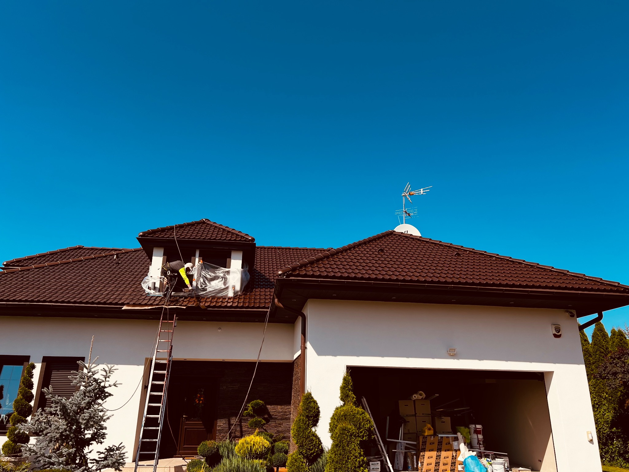 Pracownik zabezpiecza okno folią na dachu domu z brązową dachówką. Przy elewacji stoi drabina. Błękitne niebo w tle. Przygotowania do prac na wysokości.