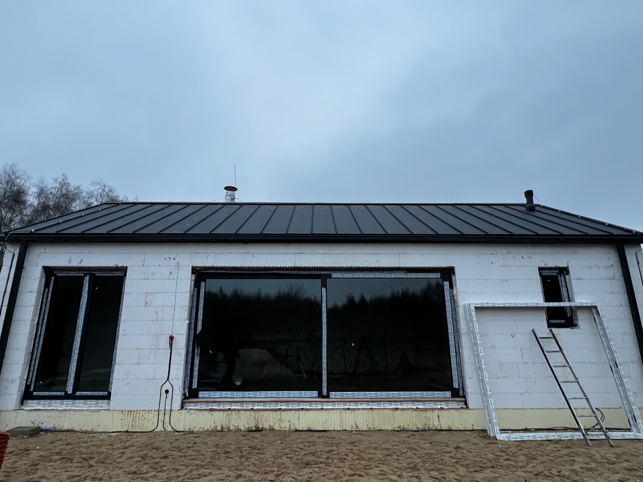 Dom w trakcie budowy z czarnym dachem, widoczne okna i drabina oparta o ścianę. Budynek z białego bloczka, surowy stan, pochmurne niebo w tle.