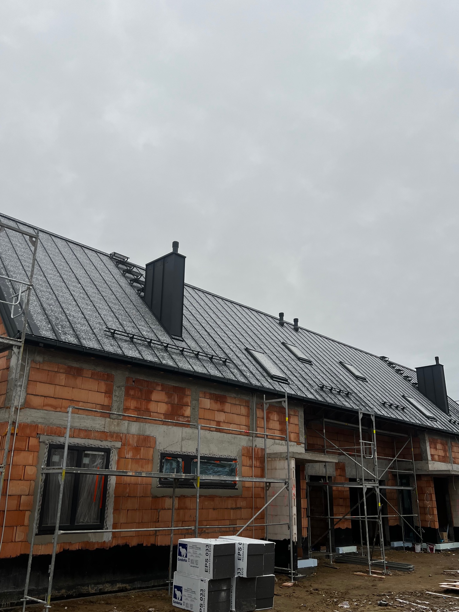 Nowy, grafitowy dach z blachy na surowym budynku z czerwonej cegły. Widoczne rusztowania i okna dachowe. Pochmurne niebo w tle.