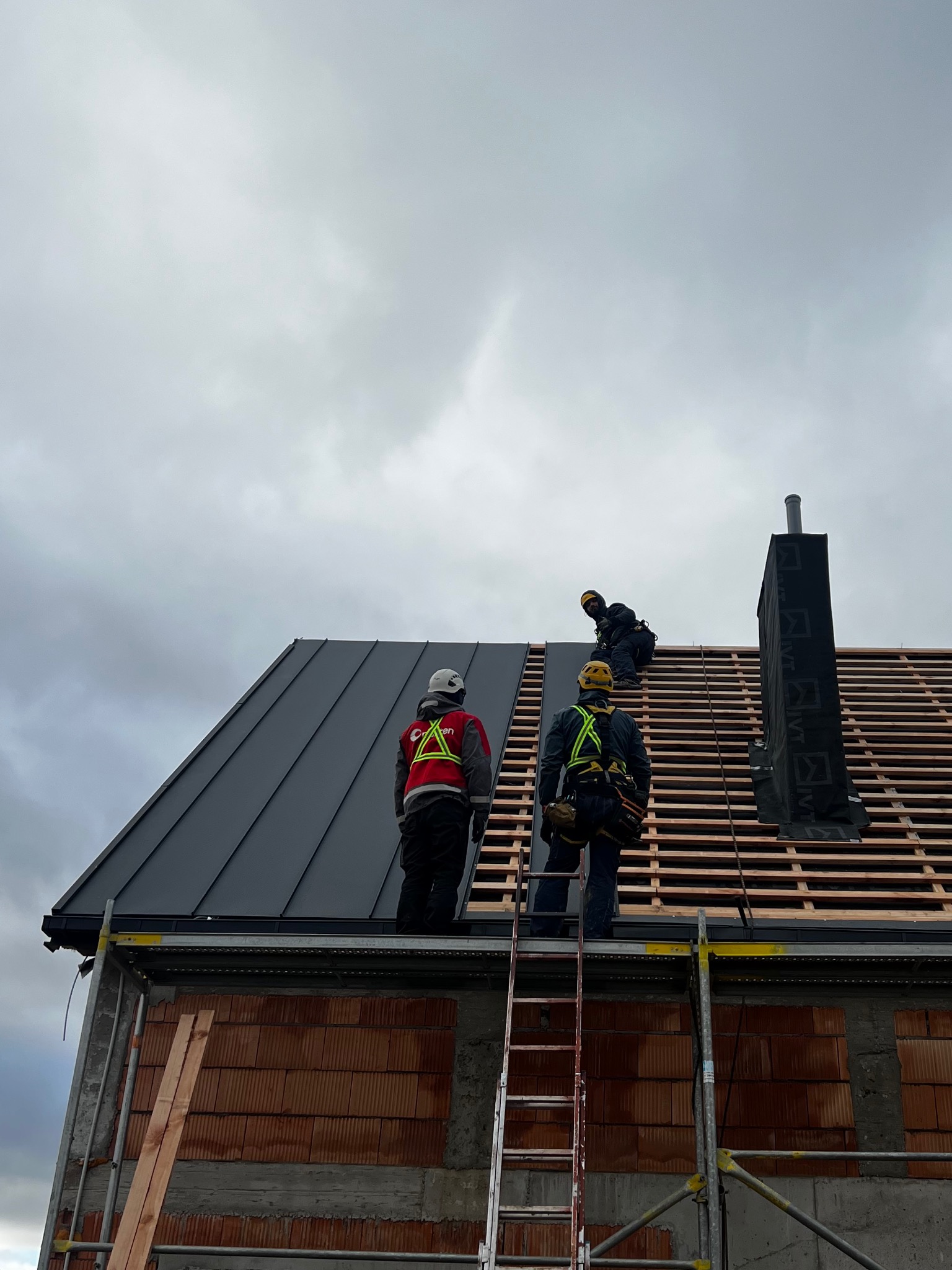 Montaż dachu z blachy na budynku w trakcie budowy. Widoczni dekarze w uprzężach, rusztowanie i komin. Część dachu pokryta blachą, reszta w konstrukcji drewnianej.