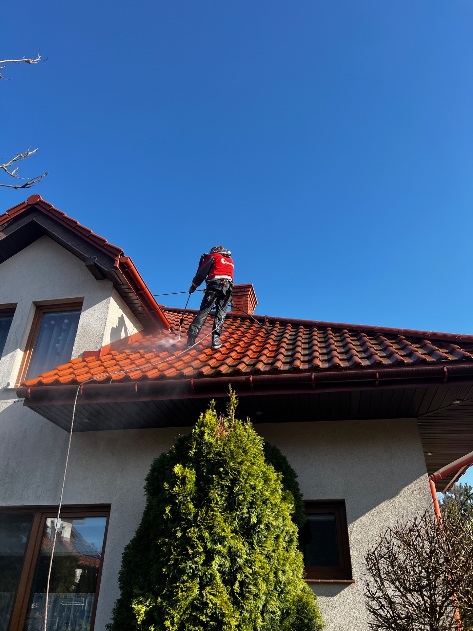 Pracownik w Łodzi czyści dach z czerwonej dachówki za pomocą myjki ciśnieniowej. Widoczny dom z jasną elewacją i błękitne niebo w tle. Prace na wysokości.