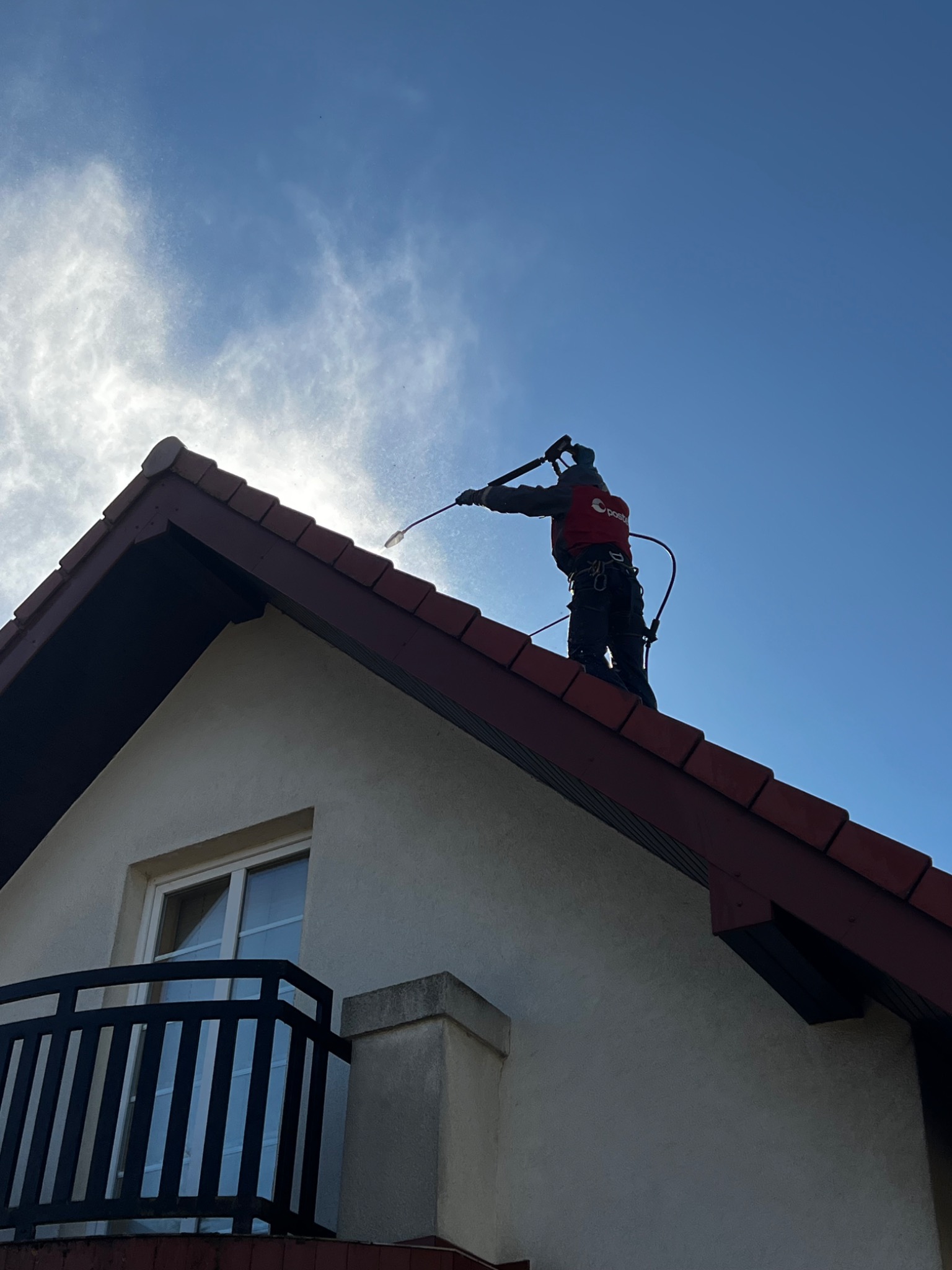Pracownik w uprzęży, z perspektywy żabiej, czyści dach domu za pomocą myjki ciśnieniowej. W tle błękitne niebo i słońce.