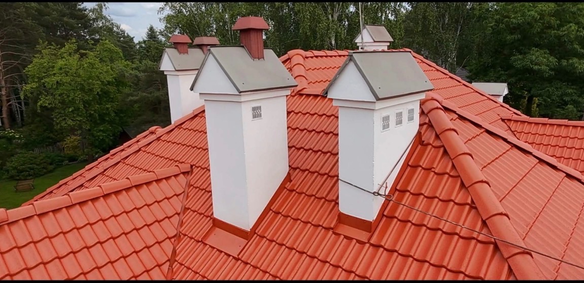 Czerwony dach z dachówki ceramicznej z białymi kominami na tle zieleni drzew. Widok z góry, prezentujący detale pokrycia i wykończenia.