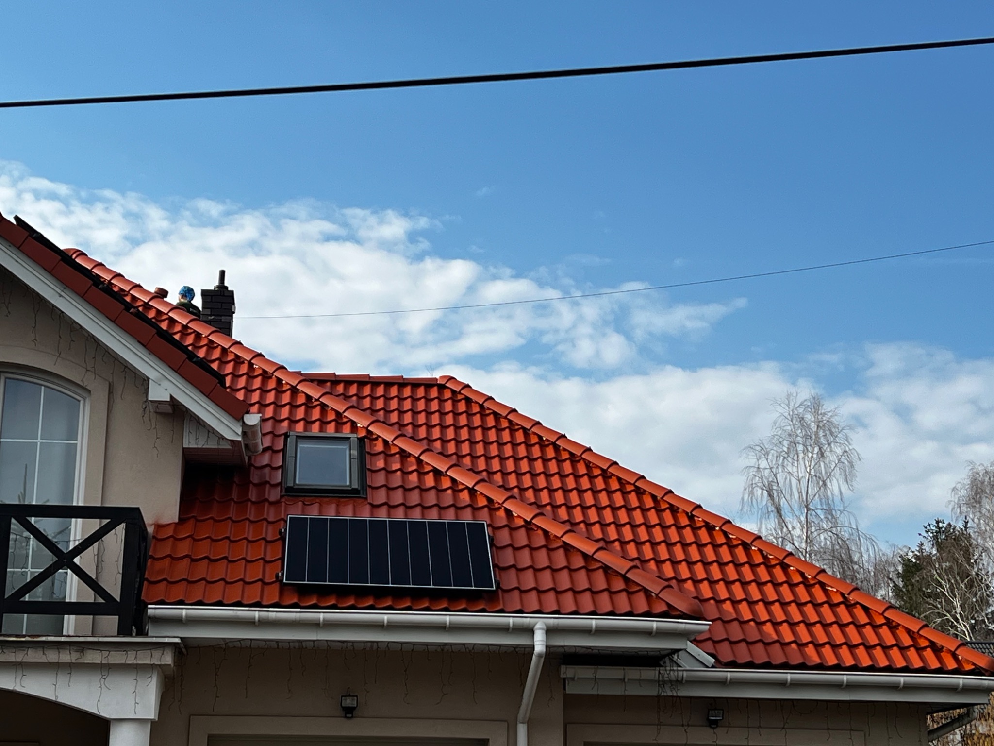 Czerwony dach z dachówki ceramicznej, okno dachowe i panel solarny. Widoczny fragment elewacji budynku i błękitne niebo z chmurami w tle.