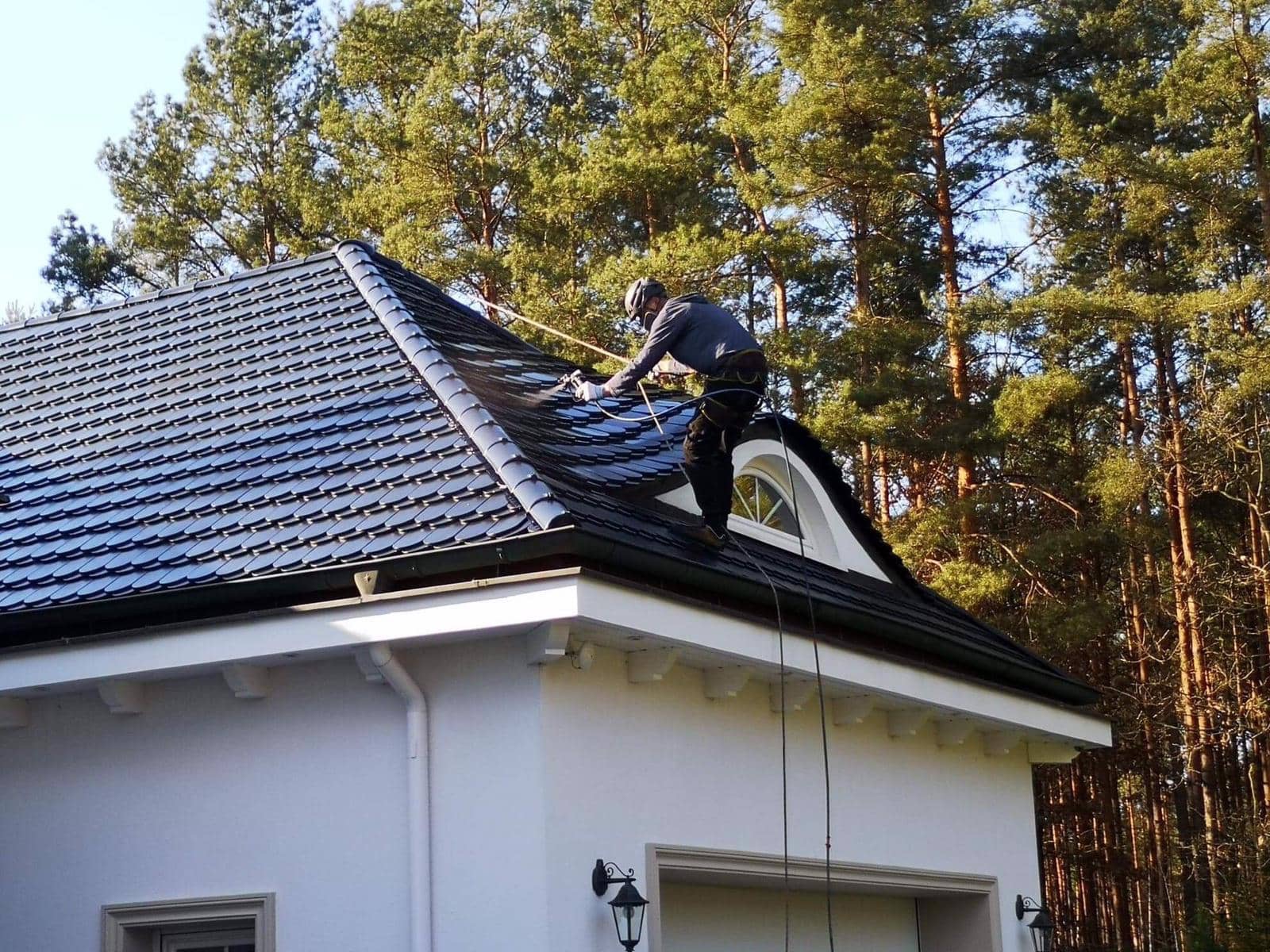 Pracownik w kasku i uprzęży myje ciemny dach domu z owalnym oknem na poddaszu, używając lancy ciśnieniowej. W tle wysokie drzewa.