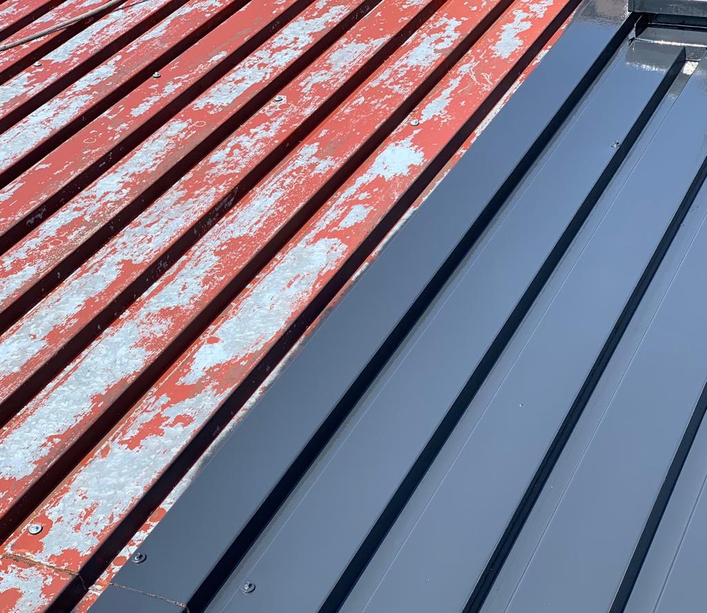 Częściowo pomalowany blaszany dach: stara, łuszcząca się czerwona farba kontrastuje z nową, gładką, szarą powłoką. Widoczne mocowania.