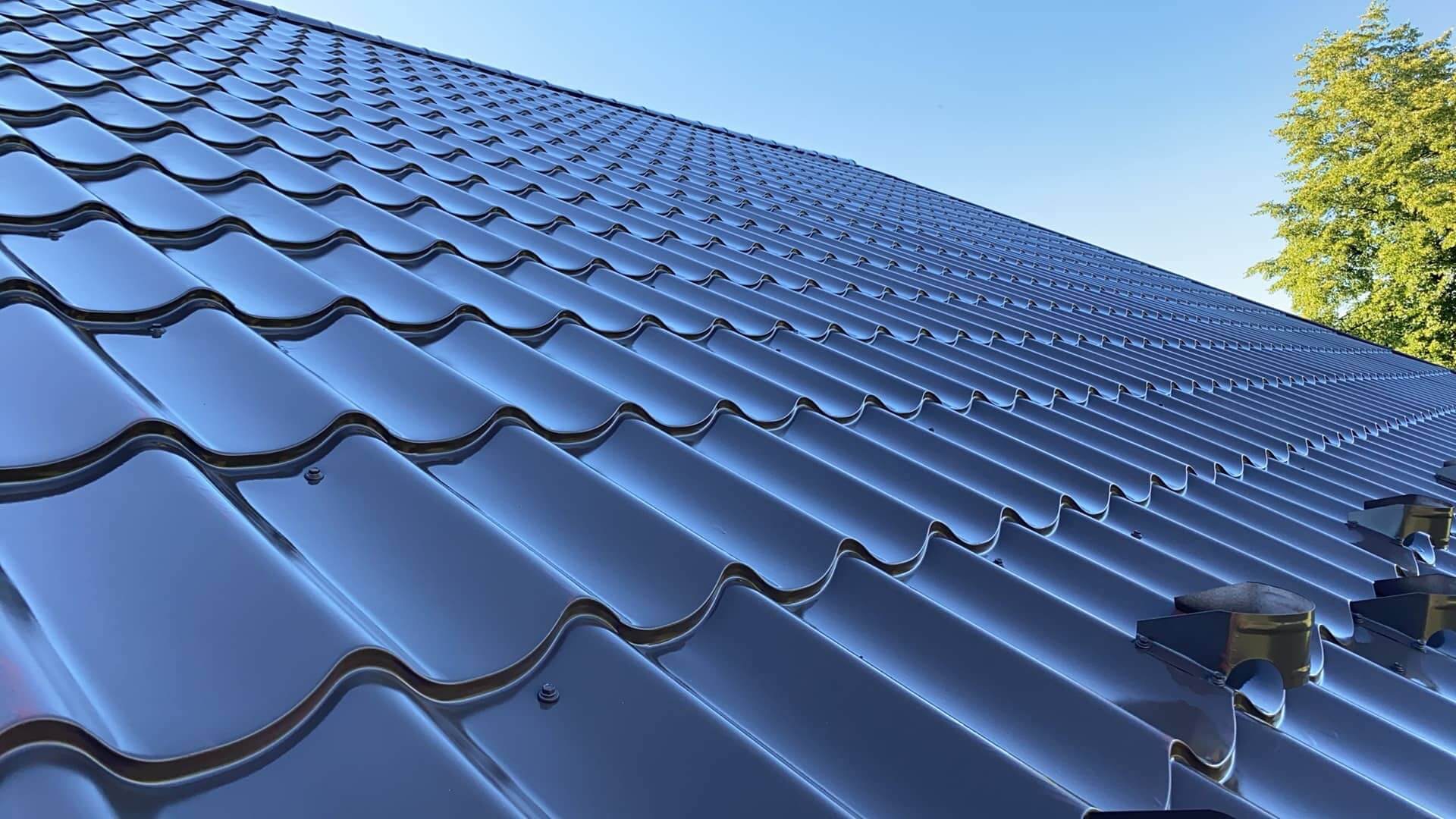 Błyszczący, ciemny dach z blachy trapezowej pod błękitnym niebem, widok z dołu. Widoczne detale łączeń i fragment drzewa w tle.