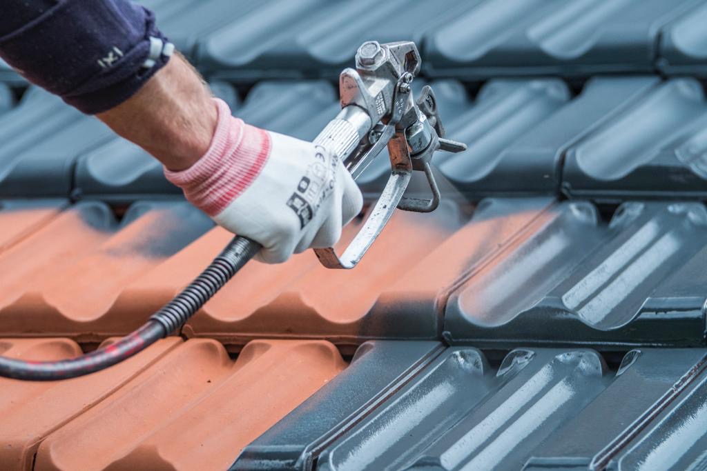 Malowanie dachówki pistoletem natryskowym. Widoczna pomarańczowa i szara dachówka. Ręka w rękawicy trzyma pistolet. Detal z bliska, ostre światło.
