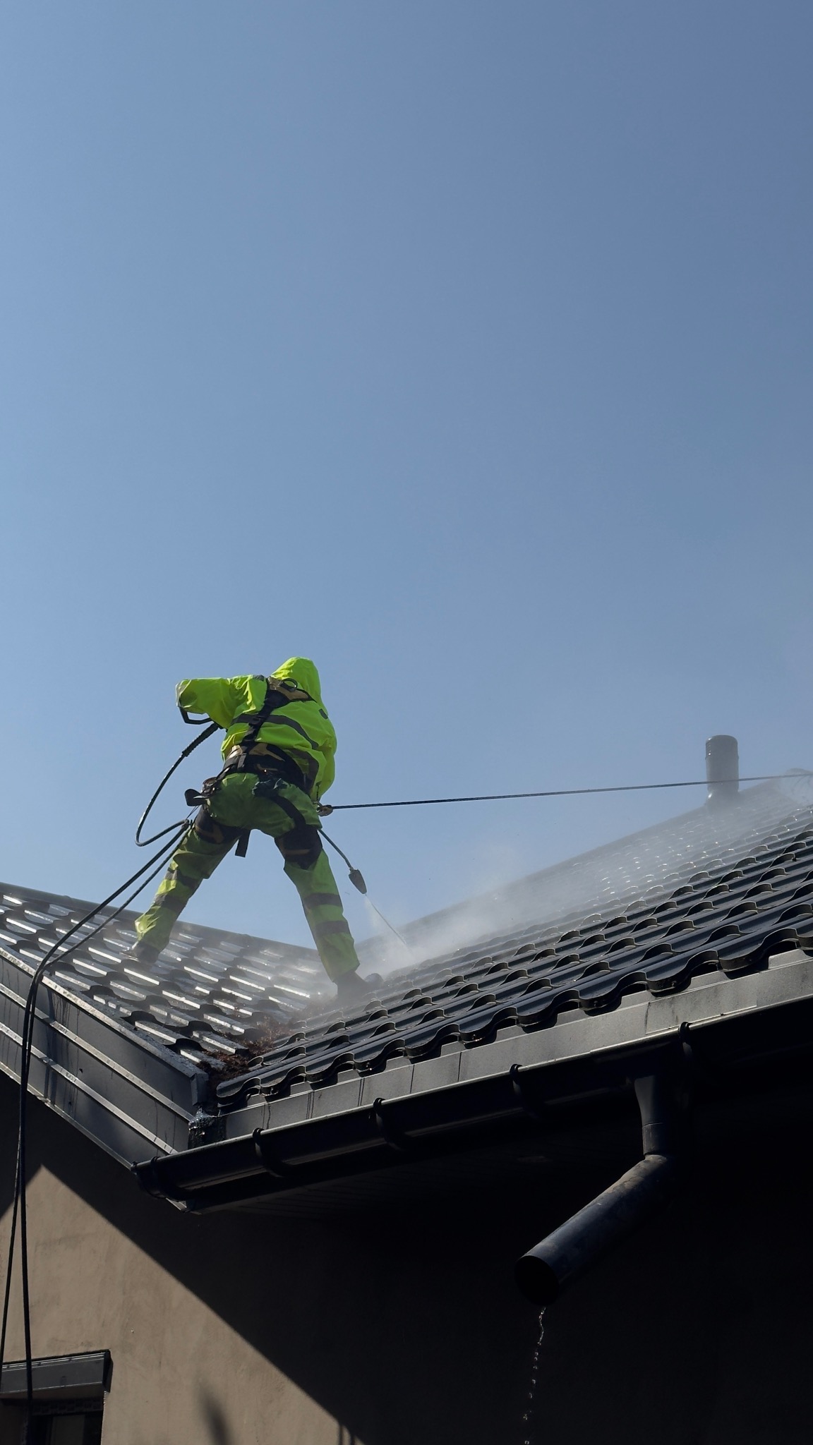 Pracownik w jaskrawozielonym kombinezonie, zabezpieczony linami, czyści dach z ciemnej dachówki za pomocą myjki ciśnieniowej. Woda spływa po dachu.