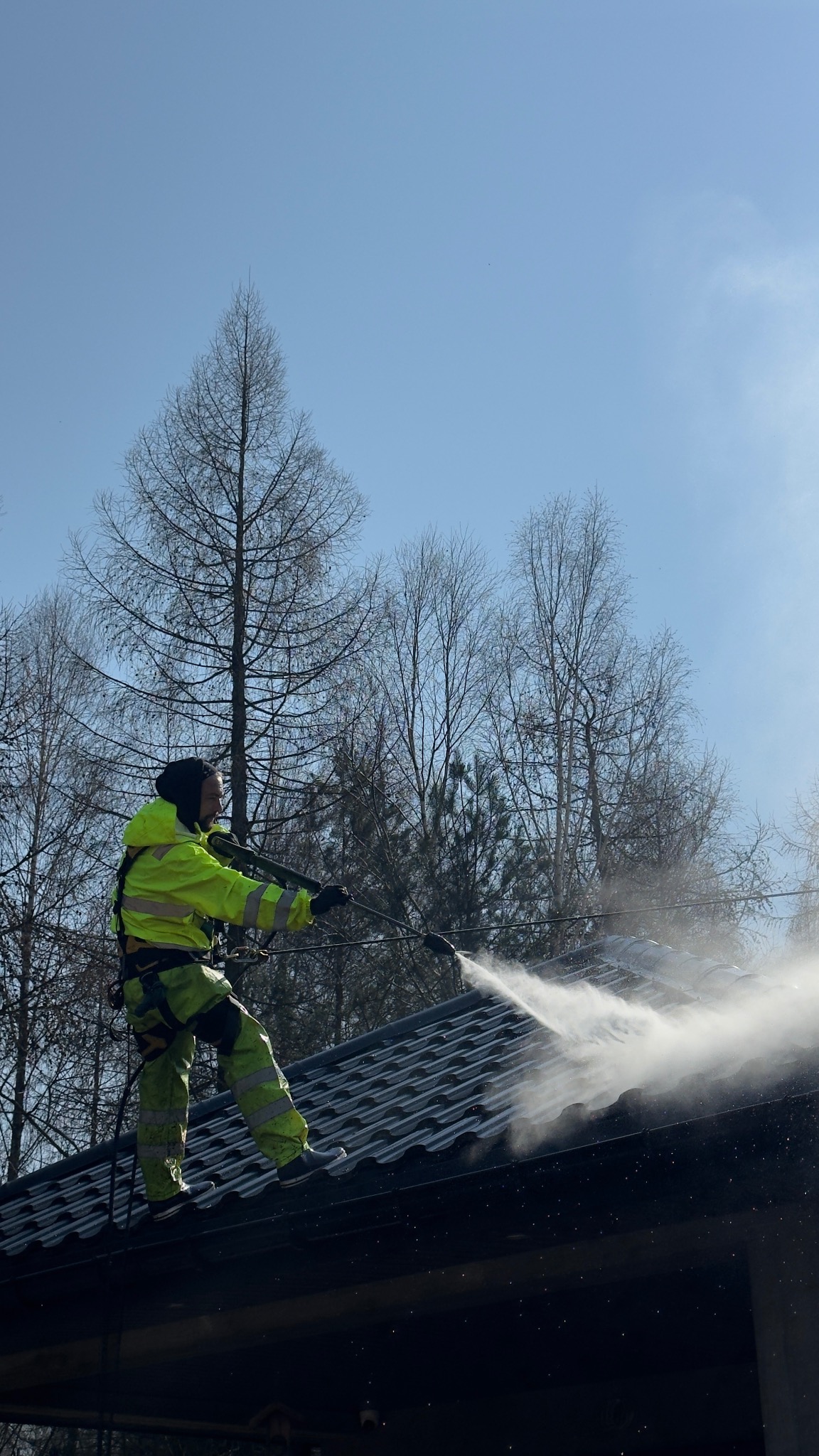 Pracownik w odblaskowym kombinezonie myje dach z metalowej dachówki za pomocą myjki ciśnieniowej. W tle błękitne niebo i drzewa bez liści.