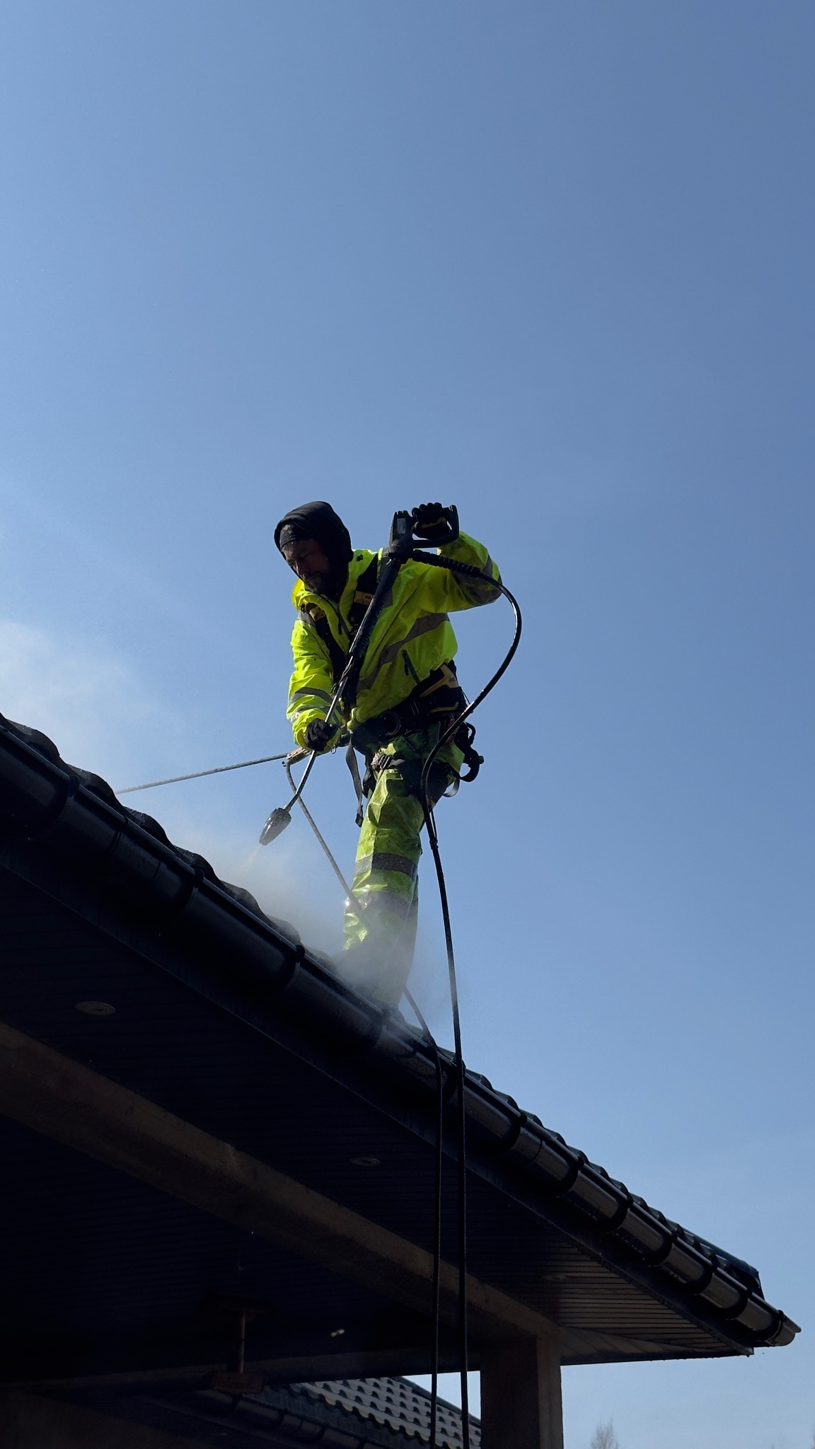 Pracownik w odzieży ochronnej czyści dach z użyciem myjki ciśnieniowej. Widoczny strumień wody i para. Ujęcie z dołu, na tle błękitnego nieba.
