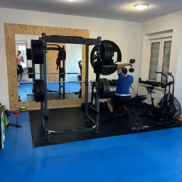 Krystian Haremza sport - Mężczyzna w niebieskiej koszulce ćwiczy z hantlami na ławce w siłowni, w tle klatka do ćwiczeń z obciążeniem, rowerek treningowy i lustro z odbiciem osoby.