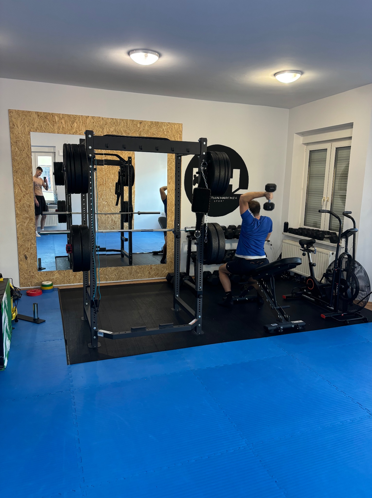 Mężczyzna w niebieskiej koszulce ćwiczy z hantlami na ławce w siłowni, w tle klatka do ćwiczeń z obciążeniem, rowerek treningowy i lustro z odbiciem osoby.