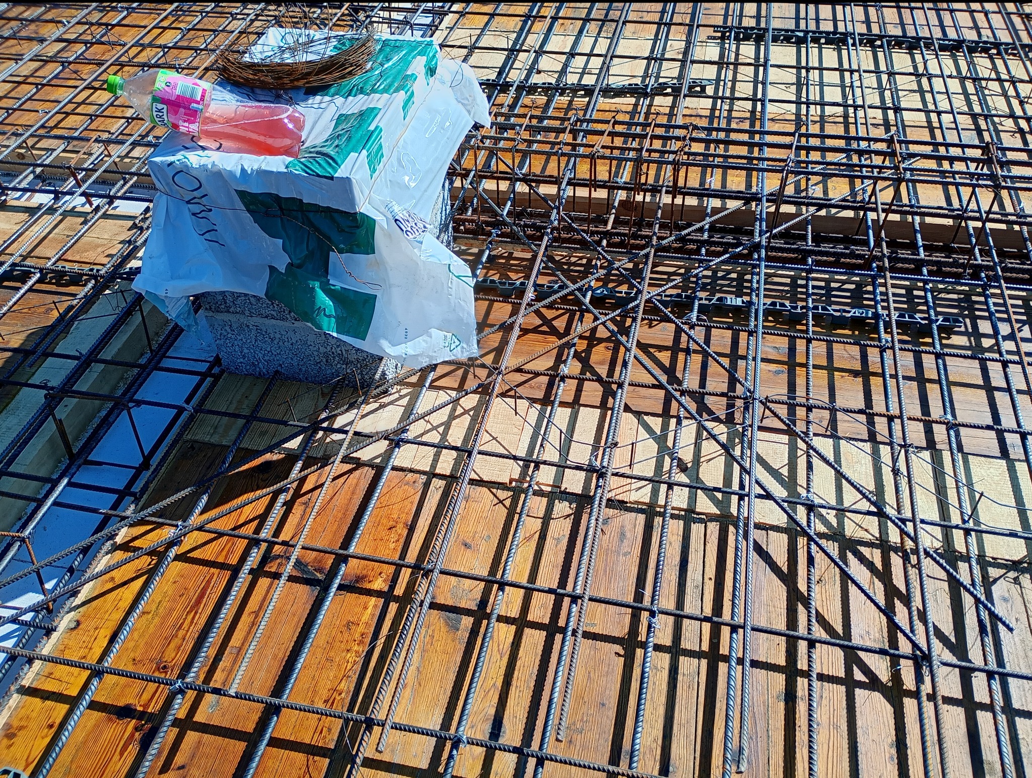 Zbrojenie stropu na deskowaniu, widok z góry. Na zbrojeniu leży worek z zaprawą i napój. Elementy konstrukcyjne w pełnym słońcu rzucają wyraźny cień.