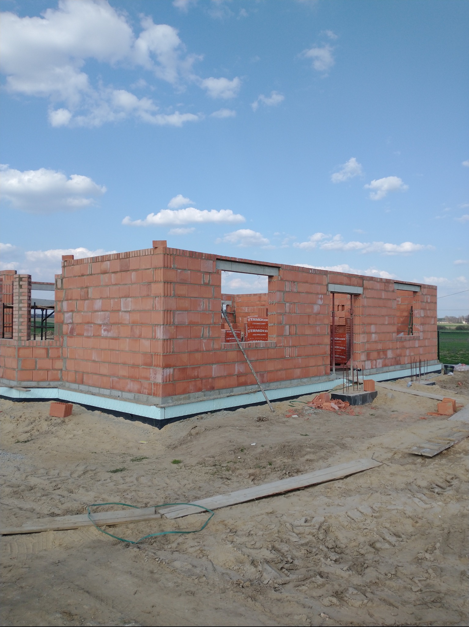 Wznoszenie ścian z czerwonej cegły na budowie domu w Wolborzu. Widoczne otwory okienne i drzwiowe, częściowo ułożone cegły. Fundamenty zabezpieczone styropianem.