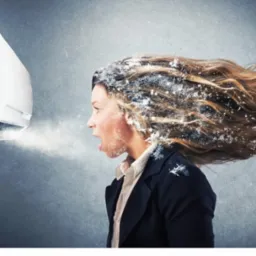 Kobieta w garniturze z otwartymi ustami, stojąca przed klimatyzatorem, z włosami i twarzą pokrytą imitacją szronu, sugerującą bardzo niską temperaturę.