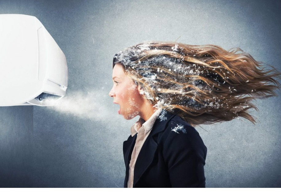 Kobieta w garniturze z otwartymi ustami, stojąca przed klimatyzatorem, z włosami i twarzą pokrytą imitacją szronu, sugerującą bardzo niską temperaturę.