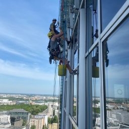 CELMAR Prace Wysokościowe Maciej Celiński - Dwóch alpinistów w kaskach myje okna wieżowca, używając lin i wiadra, z widokiem na panoramę miasta w tle.