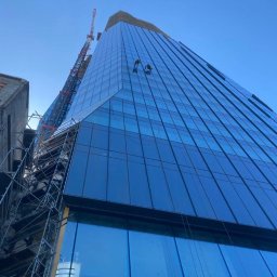 CELMAR Prace Wysokościowe Maciej Celiński - Dwóch pracowników na wysokościach, myjących szklaną fasadę nowoczesnego biurowca, widok z dołu, z widocznym rusztowaniem i dźwigiem w tle.