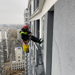 CELMAR Prace Wysokościowe Maciej Celiński - Pracownik w kasku i uprzęży, zawieszony na linie, czyści okno na wysokości w nowoczesnym budynku; w tle widok na miasto i inne budynki.