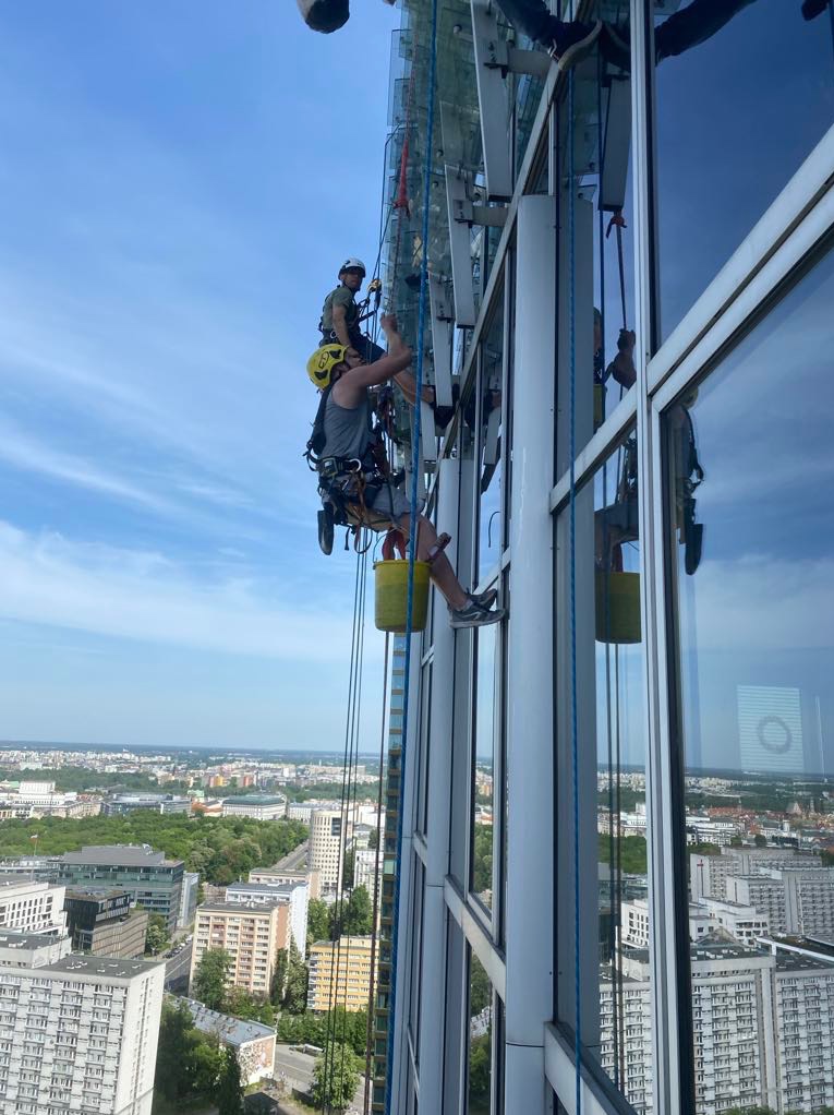 Dwóch alpinistów w kaskach myje okna wieżowca, używając lin i wiadra, z widokiem na panoramę miasta w tle.