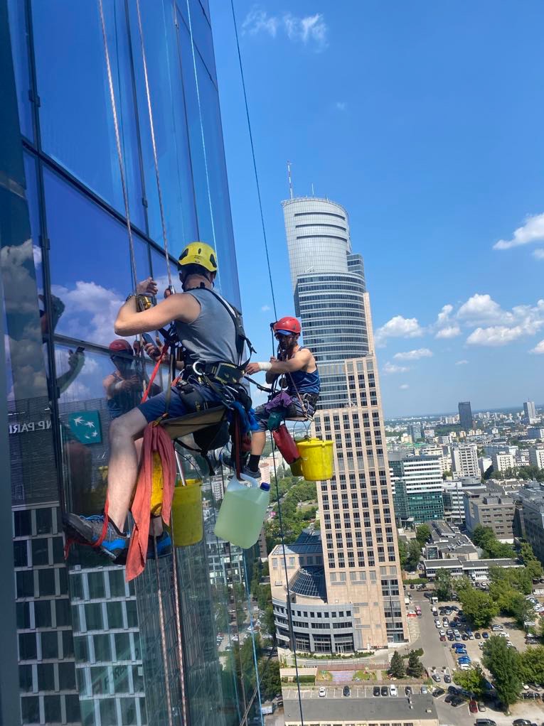 Dwóch alpinistów przemysłowych zawieszonych na linach czyści szklaną fasadę wieżowca, widok na panoramę miasta w tle.