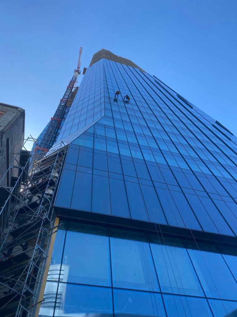 Dwóch pracowników na wysokościach, myjących szklaną fasadę nowoczesnego biurowca, widok z dołu, z widocznym rusztowaniem i dźwigiem w tle.