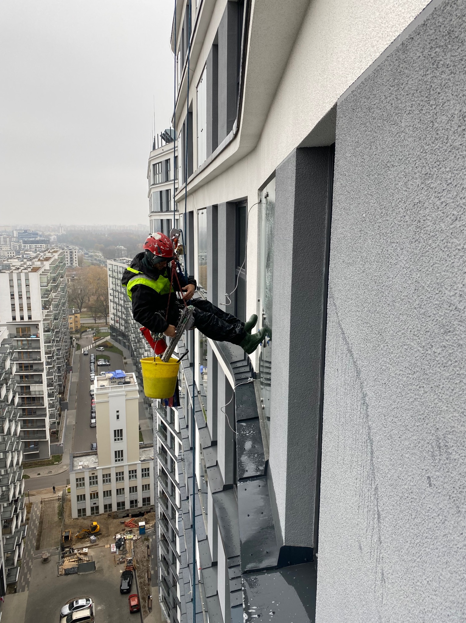 Pracownik w kasku i uprzęży, zawieszony na linie, czyści okno na wysokości w nowoczesnym budynku; w tle widok na miasto i inne budynki.