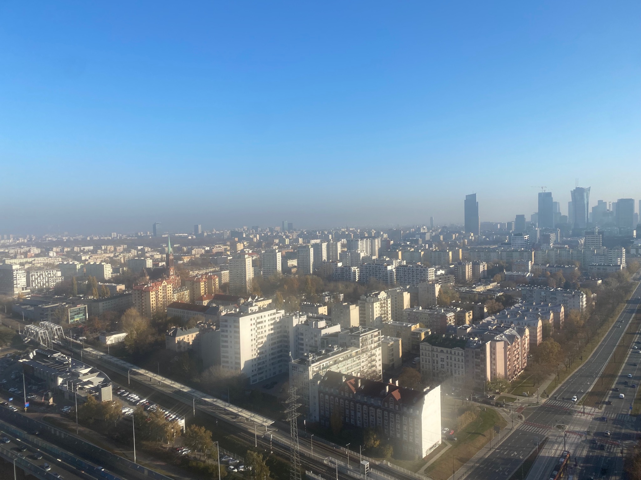 Panoramiczny widok na miasto w lekkiej mgle, z widocznymi budynkami mieszkalnymi, biurowcami i drogami, sfotografowany z dużej wysokości.