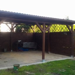 Sebastian Skrentny - Zadaszona, drewniana konstrukcja z kratownicowymi ściankami, prawdopodobnie wiata lub garaż, z widocznym fragmentem dachu i rynny, na betonowej wylewce, z elementami wyposażenia wewnątrz...