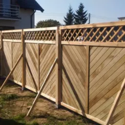 Nowy drewniany płot z ukośnymi deskami i kratką na górze, podparty tymczasowymi drewnianymi wspornikami, na tle domu jednorodzinnego.