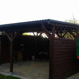 Sebastian Skrentny - Zadaszona konstrukcja drewniana z kratownicami i rynną, przylegająca do drewnianego ogrodzenia, z fragmentem zielonej przyczepy w tle.