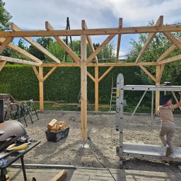 Sebastian Skrentny - Konstrukcja altany ogrodowej w trakcie budowy: drewniane słupy i belki, dziecko na podnośniku, narzędzia i materiały budowlane w tle.