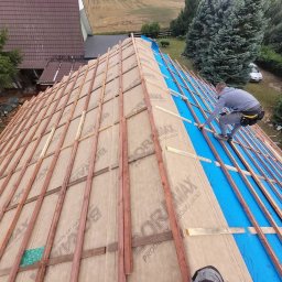 Sebastian Skrentny - Dekarz montuje kontrłaty na membranie dachowej, widok z góry na rozległy dach w trakcie prac, w tle dom i zieleń.