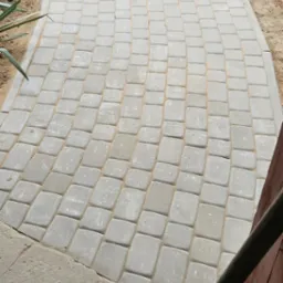 Ścieżka z szarej kostki brukowej ułożona łukiem, widoczny fragment szczotki z pomarańczową taśmą na trzonku oraz roślinność w tle.