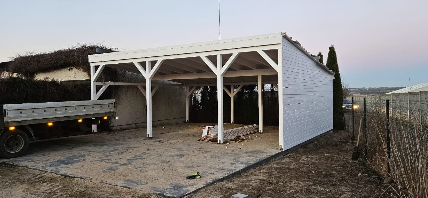 Biała konstrukcja z drewna, prawdopodobnie wiata garażowa, z elementami w stylu pergoli, częściowo zabudowana z jednej strony deskami, obok zaparkowany samochód dostawczy, widoczne materiały...