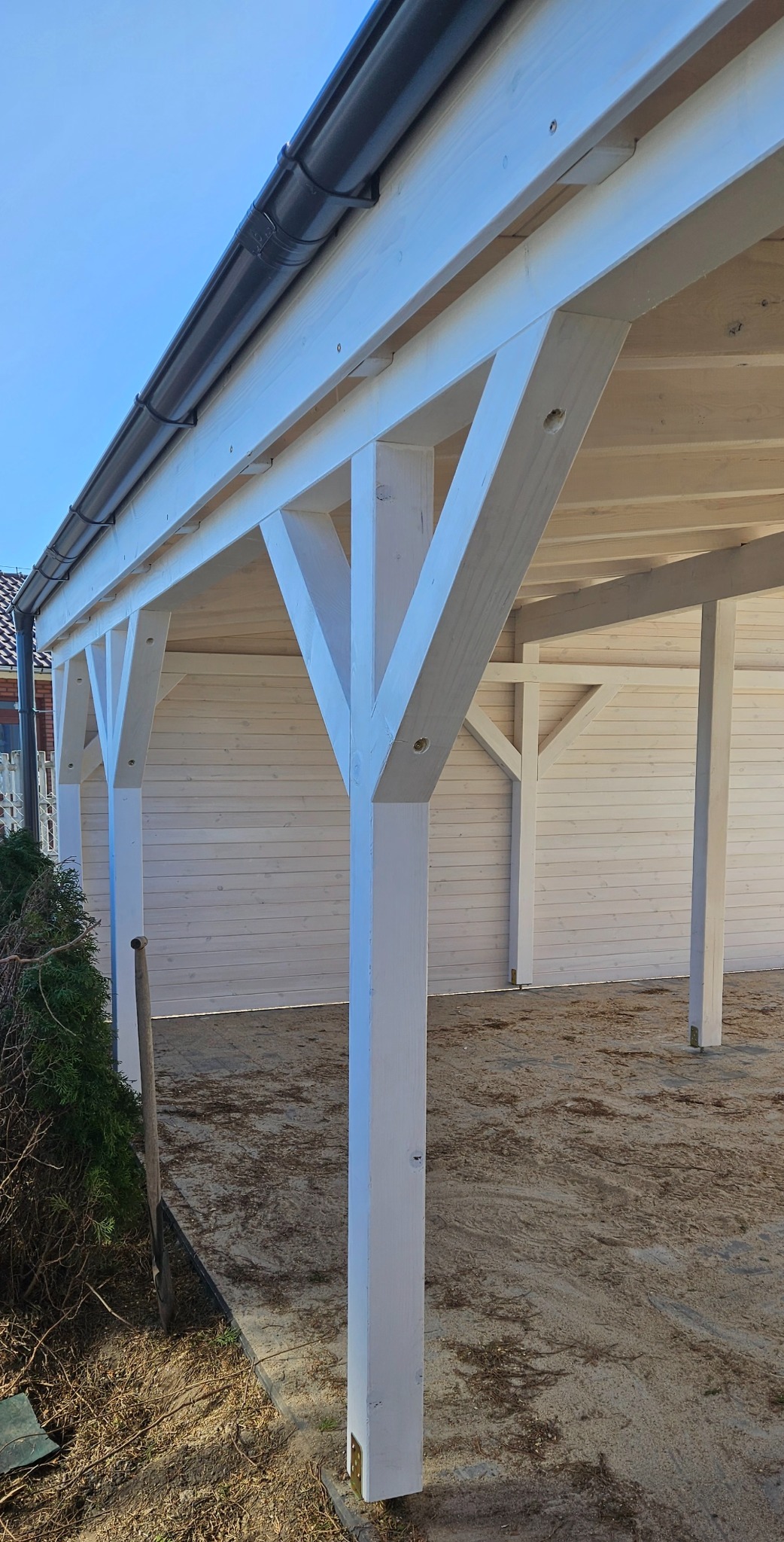 Biała, drewniana altana z szarą rynną, widok z zewnątrz na konstrukcję nośną i poszycie ścian, z narzędziem ogrodniczym opartym o jeden ze słupów.