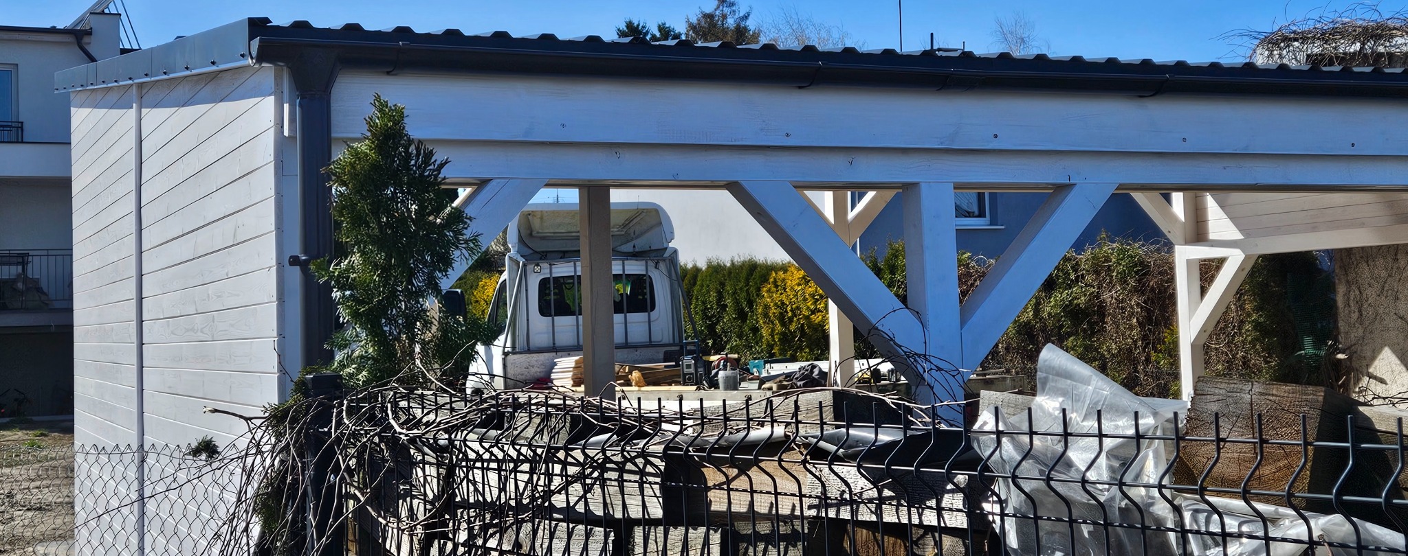 Biała drewniana konstrukcja z widocznymi elementami dachu i rynnami, częściowo zasłonięta przez metalowe ogrodzenie i splątane gałęzie, z zaparkowanym pojazdem dostawczym w tle.