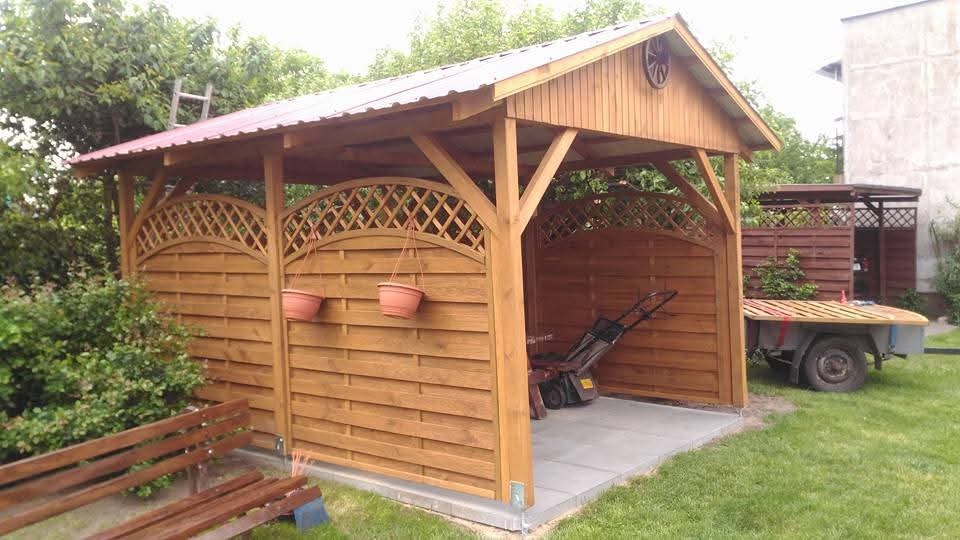 Zadaszona konstrukcja z drewna z dekoracyjnymi panelami i wiszącymi doniczkami, widoczny wózek i przyczepka w tle.