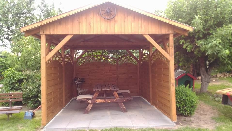 Drewniana altana z dekoracyjnym kołem na szczycie, wyposażona w stół piknikowy i kratowe panele boczne, otoczona zielenią ogrodu.