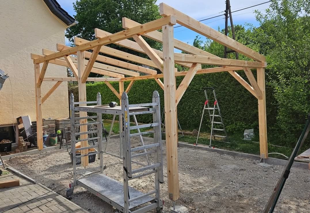 Konstrukcja z jasnego drewna, prawdopodobnie pergola, w trakcie montażu. Widoczne rusztowanie, drabina i teren przygotowany pod budowę.