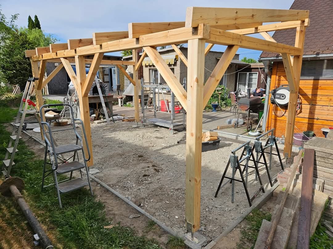 Konstrukcja drewnianej pergoli w trakcie budowy, widoczne słupy i belkowanie, teren przygotowany, narzędzia pracy wokół.
