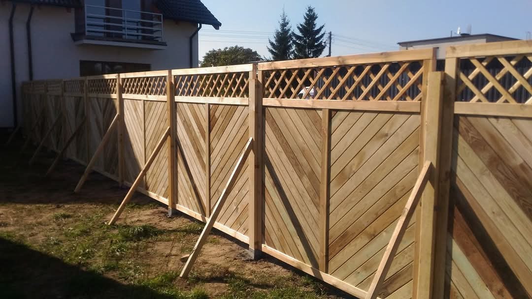 Nowy drewniany płot z ukośnymi deskami i kratką na górze, podparty tymczasowymi drewnianymi wspornikami, na tle domu jednorodzinnego.