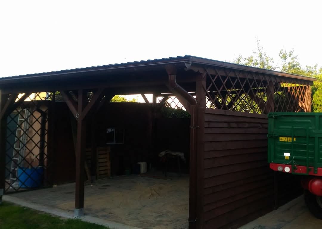 Zadaszona konstrukcja drewniana z kratownicami i rynną, przylegająca do drewnianego ogrodzenia, z fragmentem zielonej przyczepy w tle.