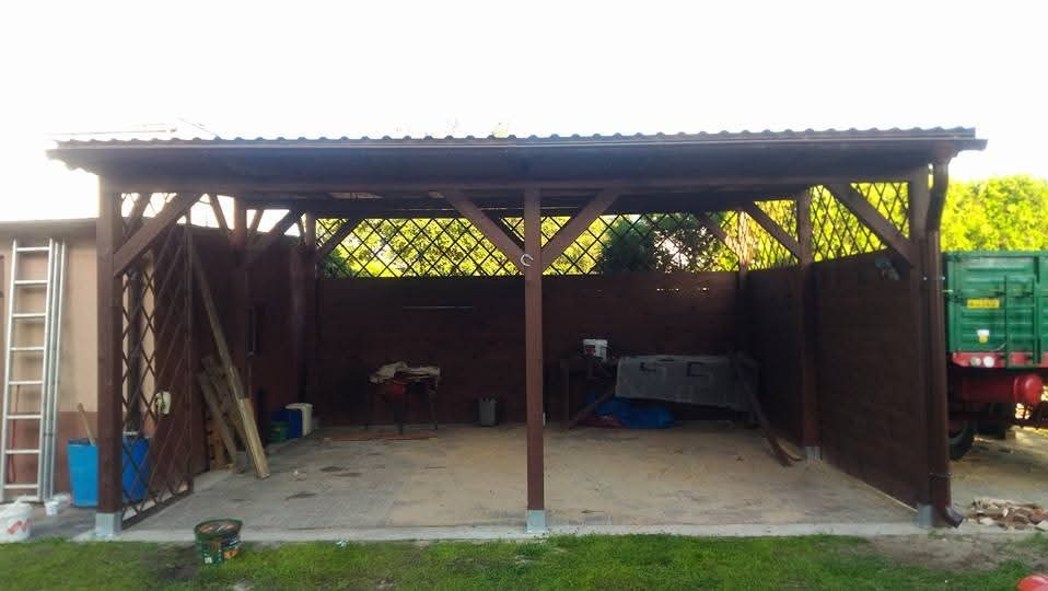 Drewniana konstrukcja z zadaszeniem i ażurowymi ścianami, z widocznym fragmentem zielonej ciężarówki w tle, na jednym ze słupów przymocowana podkowa.