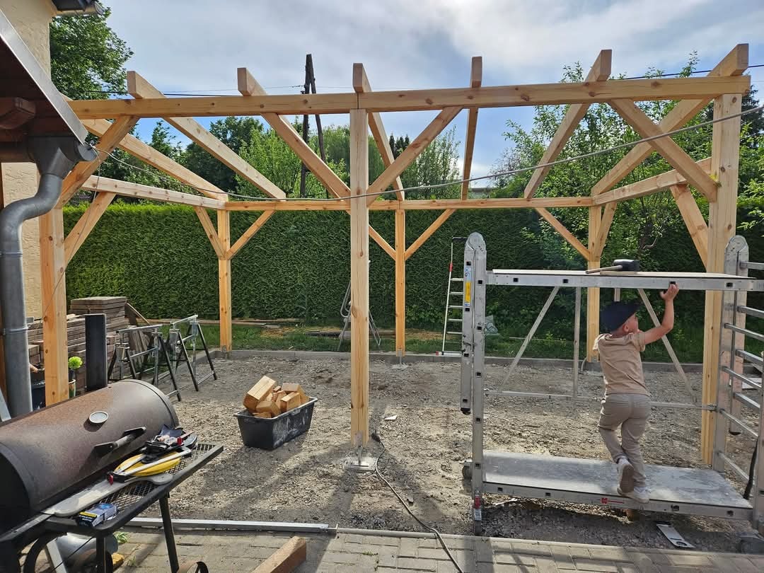 Konstrukcja altany ogrodowej w trakcie budowy: drewniane słupy i belki, dziecko na podnośniku, narzędzia i materiały budowlane w tle.